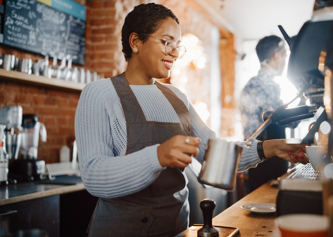 Beautiful Latin American Female Barista with Short Hair and Glasses is Making a Cup of Tasty Cappuccino in Coffee Shop Bar. Portrait of Happy Employee Behind Cozy Loft-Style Cafe Counter.
